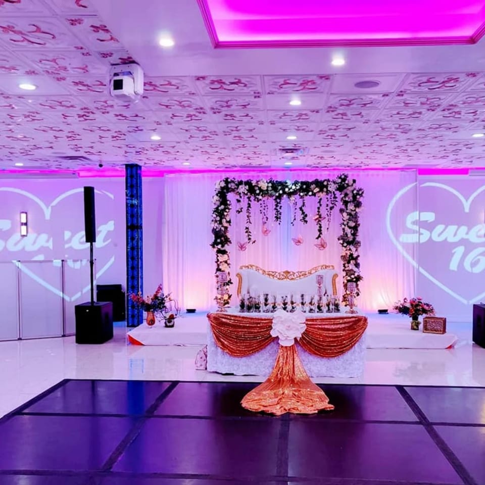 A vibrant event hall with a decorated stage, white seating, and "Sweet 16" projected on the wall.