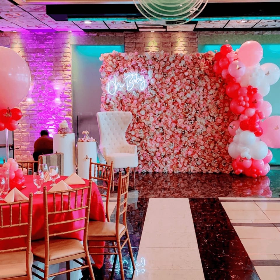 A Guru Palace event space decorated for a baby shower with pink and red balloons, a floral wall, and a neon "Oh Baby" sign.