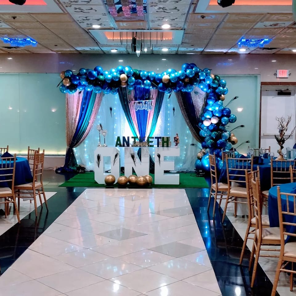A decorated event space with blue and gold balloons, a "ONE" sign, and dining tables.
