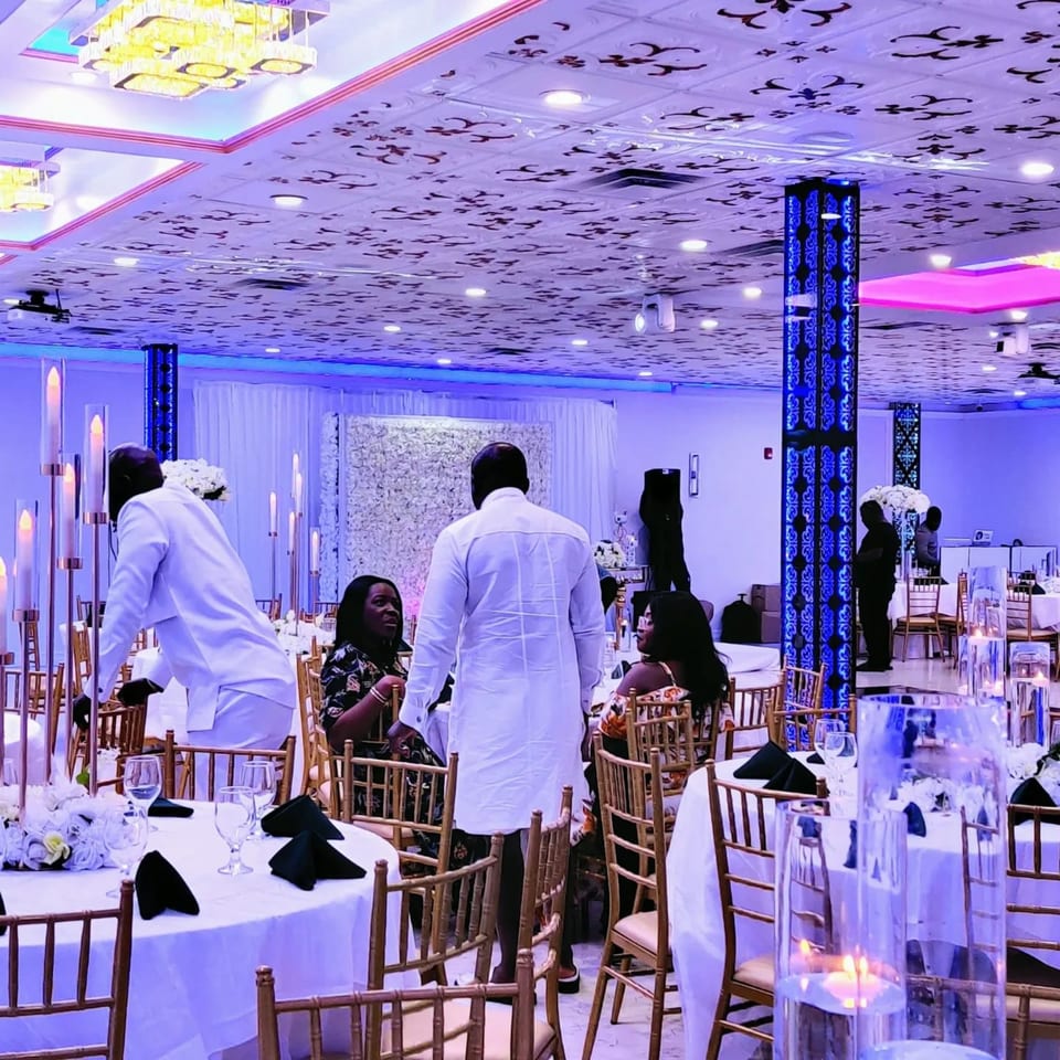 A large, elegant event space with white tables, gold chairs, and people mingling under blue lighting.